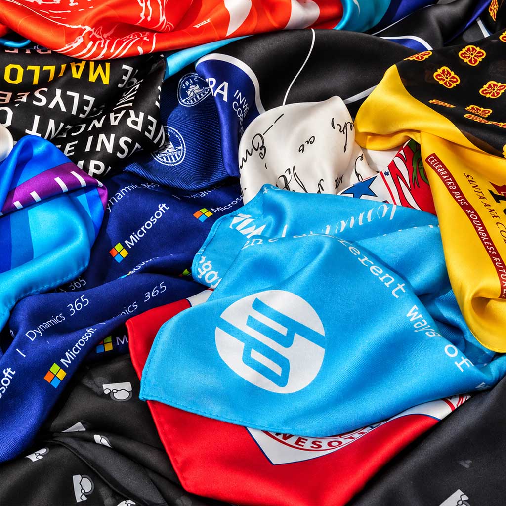 A close-up pile of colorful silk and satin scarves with printed logos and graphics, including Microsoft branding, black, blue, red, yellow, white, and orange sections. These scarves were designed and manufactured by anne touraine USA
