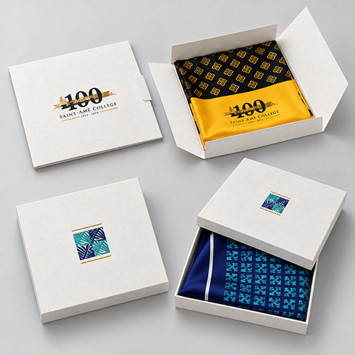 scarf envelopes and scarf boxes are arranged on a light gray background, each containing a folded patterned scarf. The open envelope shows a logoed black scarf with gold geometric patterns; the open box shows a blue scarf with a blue geometric pattern; the closed box shows the front logo design.